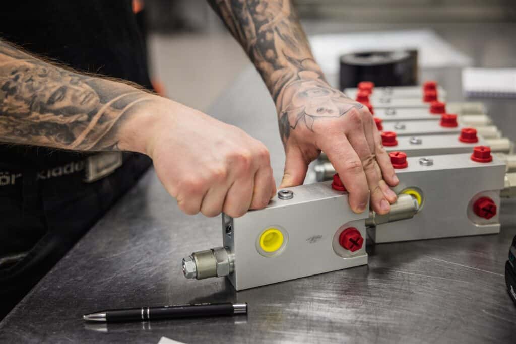 Tattooed person assembling precision metal industrial components on a workbench.